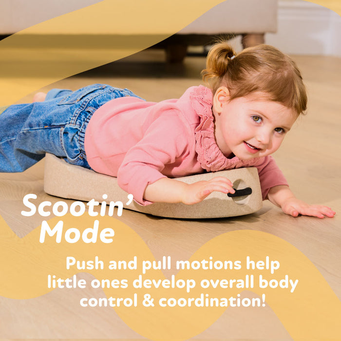 A young child smiles while lying on their stomach atop the Rock 'N Ride | Pink 3-in-1 ride-on toy on a wooden floor. Text: Scootin Mode. Push and pull actions help develop body control and coordination!.