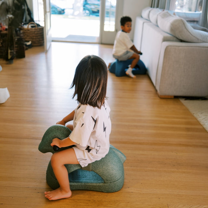 Two young children play indoors on Rock 'N Ride | Sage 3-in-1 ride-on toys. One child faces away from the camera, while the other rides near a gray couch. Sunlight shines through large windows behind them.