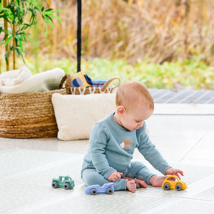 A baby in blue sits on the floor holding the Nuby Silicone Vehicle Teether Toy | Car and playing with three toy cars—yellow, blue, and green. Behind are a woven basket, cream cushion, and greenery outside.