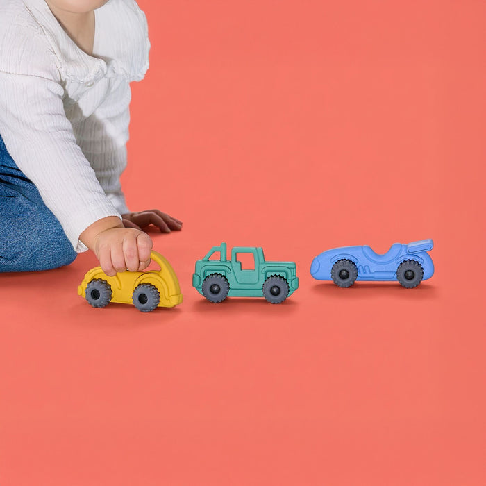 A child in a white shirt and blue pants pushes a yellow toy car beside green and blue vehicles on a coral floor, with the Nuby Silicone Vehicle Teether Toy | Truck nearby for soothing playtime breaks.