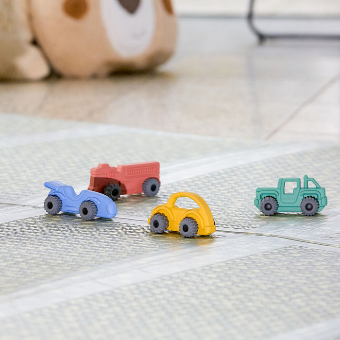 A Nuby Silicone Vehicle Teether Toy in the shape of a red truck sits on a patterned floor mat alongside other colorful toy vehicles, while a plush bear-faced toy is visible in the background.