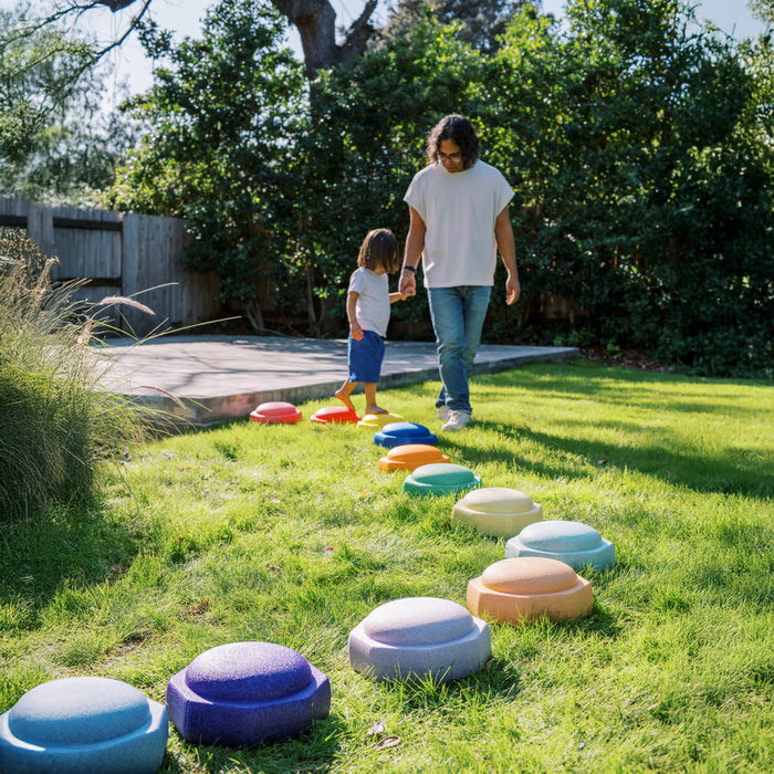 An adult and child walk hand in hand across StepPebbles and FREE Balance Board | Pastel, colorful foam stepping stones arranged on grass, perfect for indoor or outdoor play to help kids develop balance and coordination.