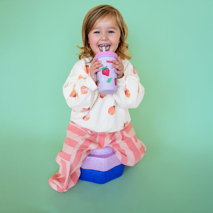 A smiling young child sits on stacked foam blocks holding the Nuby Thirsty Kids Flexstraw Stainless Steel Cup | Strawberries, wearing a white shirt with orange patterns and pink striped pants, against a mint green background.